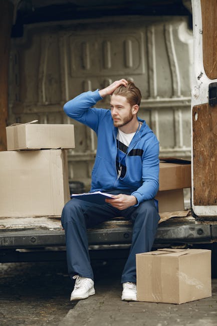 A young man with light brown hair and a beard, dressed in a blue zip-up hoodie, navy trousers, and white sneakers, is sitting on the edge of an open van located in a driveway or loading area. He is ho