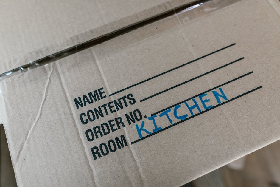Close-up image of a cardboard moving box with printed labels indicating spaces for the name, contents, order number, and room. The room label 'KITCHEN' is handwritten in blue marker. The box is partially wrapped in clear plastic, and it appears to be packed with household items as part of a home relocation process. The background suggests that the box is inside a property ready for loading onto a van, with natural lighting illuminating the scene. This image supports services related to house removals, packing, and furniture transport, highlighting organised packing practices often associated with professional moving companies like Man with Van Rainham.