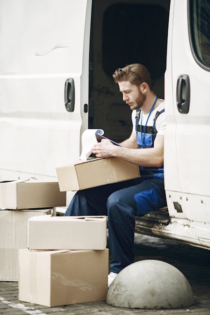 A male mover with short brown hair and a beard is sitting on the open side door of a white delivery van, inside a housing estate or driveway. He is wearing a blue and white striped t-shirt and blue work overalls, and is holding a clipboard or checklist with papers. Surrounding him are several cardboard boxes, some open and others closed, which are likely filled with household items for a home relocation. The boxes are stacked on the ground, and the mover appears to be reviewing paperwork related to the packing and moving process. The van is parked on a paved surface, with a concrete wheel stop and a curbed edge visible nearby. The environment suggests a professional moving service engaged in furniture transport and packing for a domestic move. Man with Van Rainham, a company specialising in removals, is associated with the scene, reflecting typical moving logistics and loading procedures.