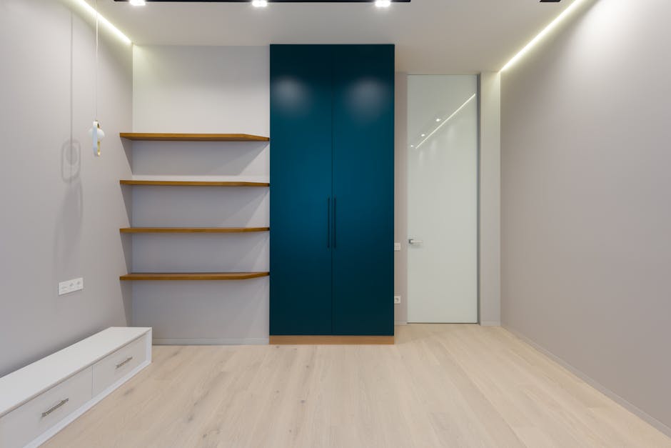 The image shows an empty interior room with minimalist design, featuring light wooden flooring and plain light grey walls. On the left side, there are three wooden floating shelves mounted on the wall above a low white storage unit with two drawers. To the right of the shelves, there is a large, colour-blocked blue wardrobe with two long vertical handles, positioned adjacent to a white door with a handle, leading to another room or hallway. The ceiling has built-in LED strip lighting along the perimeter, providing bright, even illumination. The scene depicts a clean, modern space likely intended for residential use, and the environment suggests a home undergoing a furniture removal or packing process, with no furniture or boxes present in the room itself. The image aligns with house removals or relocation services, such as those offered by Man with Van Rainham, highlighting the transition of home interior furniture and storage units during moving logistics.