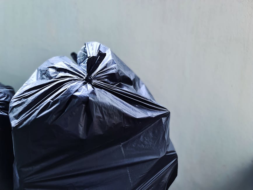 Close-up view of multiple black plastic rubbish bags filled with household waste, stacked against a plain, light-colored wall. The bags are tightly sealed, with some showing slight wrinkles and reflections due to the plastic material. They are situated near an indoor doorway, suggesting the process of clearing or preparing waste for disposal during a home relocation or packing and moving activity. The lighting is even, highlighting the texture and creases of the bags. This scene illustrates typical waste management steps involved in house removals, with the bags ready to be transported or disposed of by professional removal services such as Man with Van Rainham, especially in the context of complying with household waste duty regulations in Rainham.