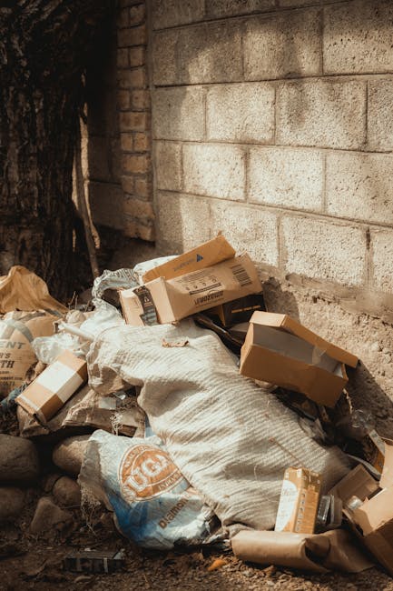 A person wearing a white shirt and an orange vest is seen placing crumpled paper and cardboard packaging into a white waste bin, positioned on a wooden surface near the edge of a table. The individual is holding the packaging with both hands, with the bin’s open top visible in the foreground. In the background, part of the person's beige trousers and torso are visible, indicating they are in an indoor setting, likely involved in packing or preparing items for disposal as part of a home relocation or household move. Nearby, there are additional packing materials, suggesting the process of packing fragile or miscellaneous items before a move. The scene is well-lit, emphasizing the packaging and the activity of sorting waste, consistent with a moving or packing service context, such as those provided by Man with Van Rainham, supporting efficient furniture transport, packing, and moving logistics.