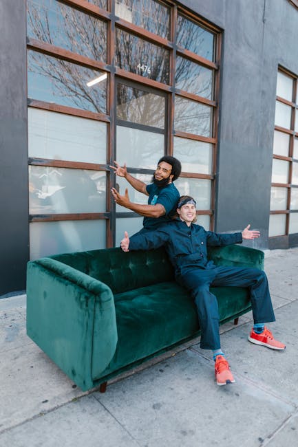 Two men are positioned outdoors in front of a commercial building with large glass windows divided by wooden frames, during daylight with natural light illuminating the scene. One man, standing on the pavement, is lifting a green velvet sofa with wooden legs, part of a home relocation process; he is wearing casual clothing, including a dark t-shirt and jeans. The other man, seated on the sofa, is wearing a dark jacket, blue pants, and red shoes, with his arms outstretched and smiling towards the camera. The scene suggests a furniture transport or packing and moving activity associated with professional removals. The building's exterior features a grey wall and visible street scene, indicating the loading or unloading area for the move undertaken by Man with Van Rainham. The image captures a moment in the furniture transport phase of a house removals service, highlighting teamwork and logistical preparation.