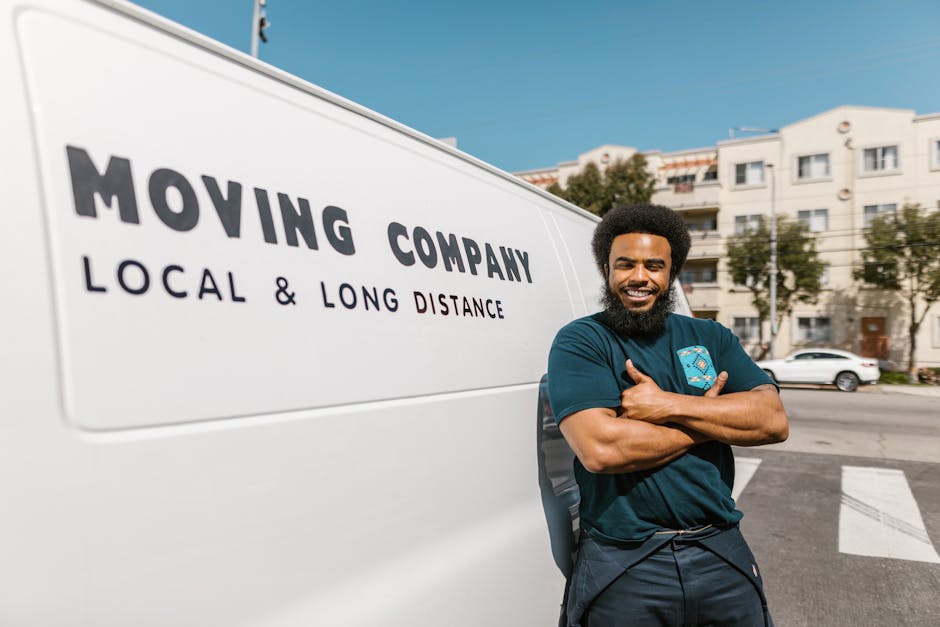 A smiling man with a full beard and curly black hair stands outside a white moving van, which displays the words 'MOVING COMPANY' and 'LOCAL & LONG DISTANCE' in bold black letters. He is wearing a dark blue t-shirt and dark pants, with his arms crossed casually as he leans against the side of the van. The background shows a row of urban residential buildings, parked cars, and a clear blue sky, indicating a bright, sunny day. The scene illustrates a home relocation or furniture transport situation, with the man representing a professional mover from Man with Van Rainham, engaged in pre-loading or post-unloading logistics near Rainham Station. The image captures the outdoor environment where packing and moving activities are taking place, including the vehicle preparation for a local or long-distance move, emphasizing the professional moving services offered.