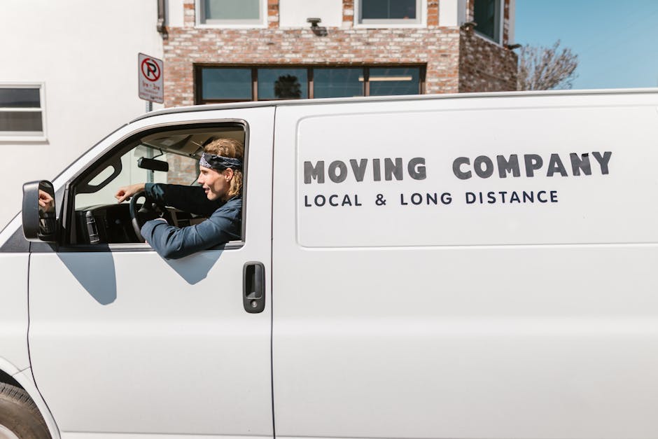 A smiling man with a full beard and curly black hair stands outside a white moving van, which displays the words 'MOVING COMPANY' and 'LOCAL & LONG DISTANCE' in bold black letters. He is wearing a dark blue t-shirt and dark pants, with his arms crossed casually as he leans against the side of the van. The background shows a row of urban residential buildings, parked cars, and a clear blue sky, indicating a bright, sunny day. The scene illustrates a home relocation or furniture transport situation, with the man representing a professional mover from Man with Van Rainham, engaged in pre-loading or post-unloading logistics near Rainham Station. The image captures the outdoor environment where packing and moving activities are taking place, including the vehicle preparation for a local or long-distance move, emphasizing the professional moving services offered.