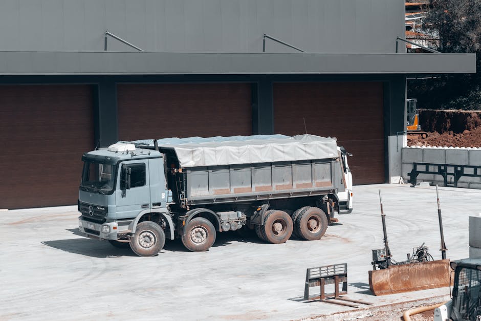 A grey flatbed truck with a covered cargo area is parked on a concrete driveway in front of a modern building with a dark brown facade and large closed garage doors. The truck is positioned close to the building, and its wheels are visible, showing some dirt and wear. Nearby, there are construction barriers, safety equipment, and a few metal poles or supports. The scene is outdoors, with lighting indicating daytime, possibly during a house or commercial relocation involving furniture transport and moving logistics. Man with Van Rainham may utilize such vehicles for their home relocation and packing and moving services, ensuring proper loading processes and vehicle positioning for efficient furniture transport and compliance with parking and loading rules in Rainham.