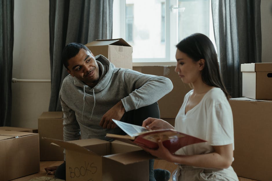 A man with a medium skin tone wearing a grey hoodie is kneeling near a collection of cardboard boxes inside a room, engaging in a conversation with a woman with a fair complexion and long dark hair, who is holding a folder or document. The cardboard boxes are of various sizes and some are labeled with words like 'books,' and are stacked on the floor near a window with light streaming in, partially covered by dark curtains. The room appears to be in the process of a home relocation, with furniture and packing materials visible, including plastic wrap around some items. The scene captures a moment of planning or discussion related to household move logistics, with the natural daylight highlighting the interior environment, which is common during a same-day move in RM13. Man with Van Rainham occasionally mentions their house removals service in Rainham, focusing on efficient and professional furniture transport and packing during scheduled or same-day moves.