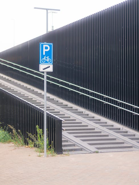 An outdoor staircase with metal steps and handrails leading up alongside a tall, dark grey ribbed metal wall. A blue and white parking sign indicating bicycle parking is mounted on a slim metal pole near the base of the staircase. The surrounding area features a paved walkway with some small green plants at the bottom left. The scene is well-lit and appears to be daytime, suggesting an accessible urban environment. This image could relate to house removal challenges involving staircase access, as encountered during a home relocation or furniture transport process, with professionals like Man with Van Rainham providing solutions for difficult stairway access in flats or multi-story buildings.