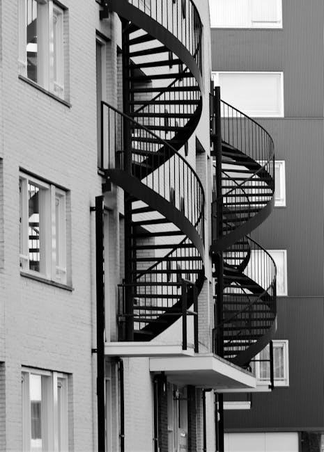 Black metal spiral staircase attached to the exterior of a modern multi-storey apartment building in Rainham, featuring open risers and curved handrails, with several landings between each turn. The staircase provides access between floors and is situated outside near windows and a small balcony area. The building’s façade includes both brick and panel cladding, with visible windows on each level. The lighting appears natural, highlighting the structure's details, and no moving or packing activities are visible in the image. This staircase may pose a challenge for home relocation or furniture transport when accessing upper floors, which is where professional removals services like those offered by Man with Van Rainham can assist in managing house and apartment moves through such staircase access issues.