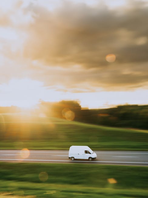A white moving van is captured in motion on a multi-lane road during sunset, with the vehicle appearing blurred due to its speed. The background features a landscape of green grass, distant trees, and a cloudy sky illuminated by the setting sun, creating a warm, golden light. This image relates to the logistics of home relocation or furniture transport, illustrating the transportation process involved in house removals, as seen in services provided by companies like Man with Van Rainham, which specialises in removals and moving arrangements near Wennington Road. The van's motion and the natural outdoor environment emphasize the importance of professional vehicle and transportation planning during packing and moving procedures.